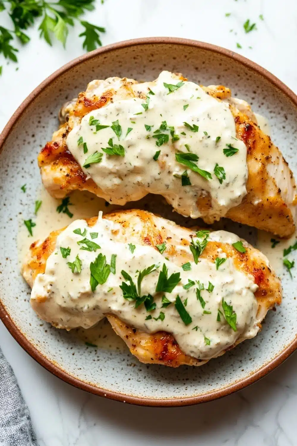 Slow cooker chicken with cream cheese - the image shows two pieces of grilled chicken breasts on a white marble countertop. the chicken breasts are golden brown and appear to be seasoned with herbs and spices. they are covered in a creamy white sauce, which is drizzled over the top. the sauce is garnished with fresh parsley leaves, adding a pop of green color to the dish. the plate is brown and has a speckled texture. a gray and white striped napkin is visible in the background.