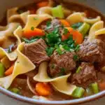 Slow cooker stew meat and noodles - the image is a close-up of a bowl of beef noodle soup. the soup is a rich brown color and appears to be thick and hearty. it is filled with chunks of beef, carrots, and celery, and is garnished with chopped parsley. the noodles are long and thin, and the soup is thick and thick. the bowl is made of ceramic and is sitting on a wooden table. the background is blurred, so the focus is on the bowl of soup.