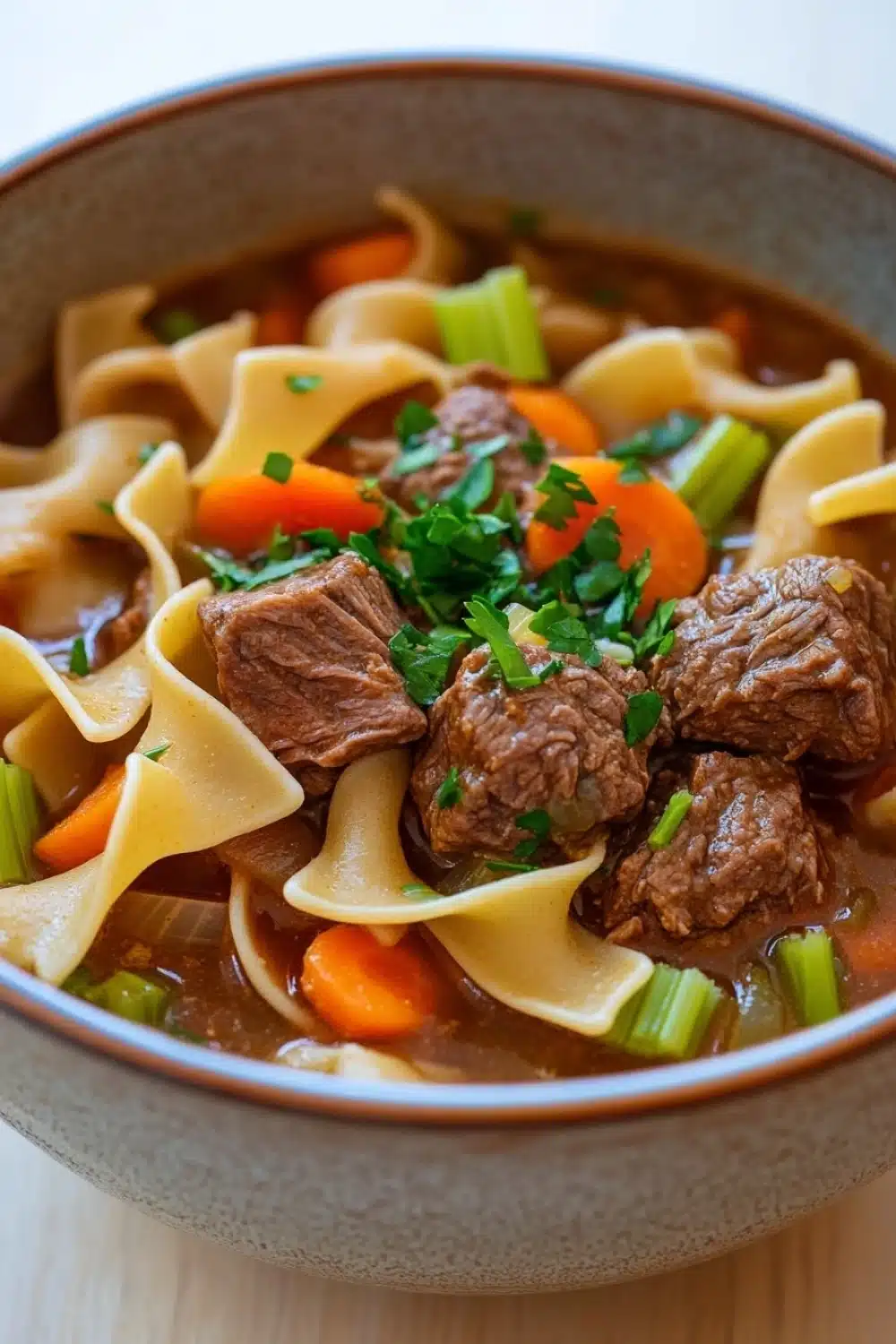 Slow cooker stew meat and noodles - the image is a close-up of a bowl of beef noodle soup. the soup is a rich brown color and appears to be thick and hearty. it is filled with chunks of beef, carrots, and celery, and is garnished with chopped parsley. the noodles are long and thin, and the soup is thick and thick. the bowl is made of ceramic and is sitting on a wooden table. the background is blurred, so the focus is on the bowl of soup.