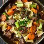 Slow cooker stew meat and noodles - the image is a close-up of a bowl of beef noodle soup. the soup is made with chunks of beef, carrots, and celery, and is garnished with chopped parsley. the bowl is made of ceramic and is sitting on a dark wooden table. the broth is a rich brown color and appears to be thick and creamy. the noodles are long and thin, and the carrots are bright orange. the celery is a bright green color and is scattered throughout the soup, adding a pop of color to the dish.