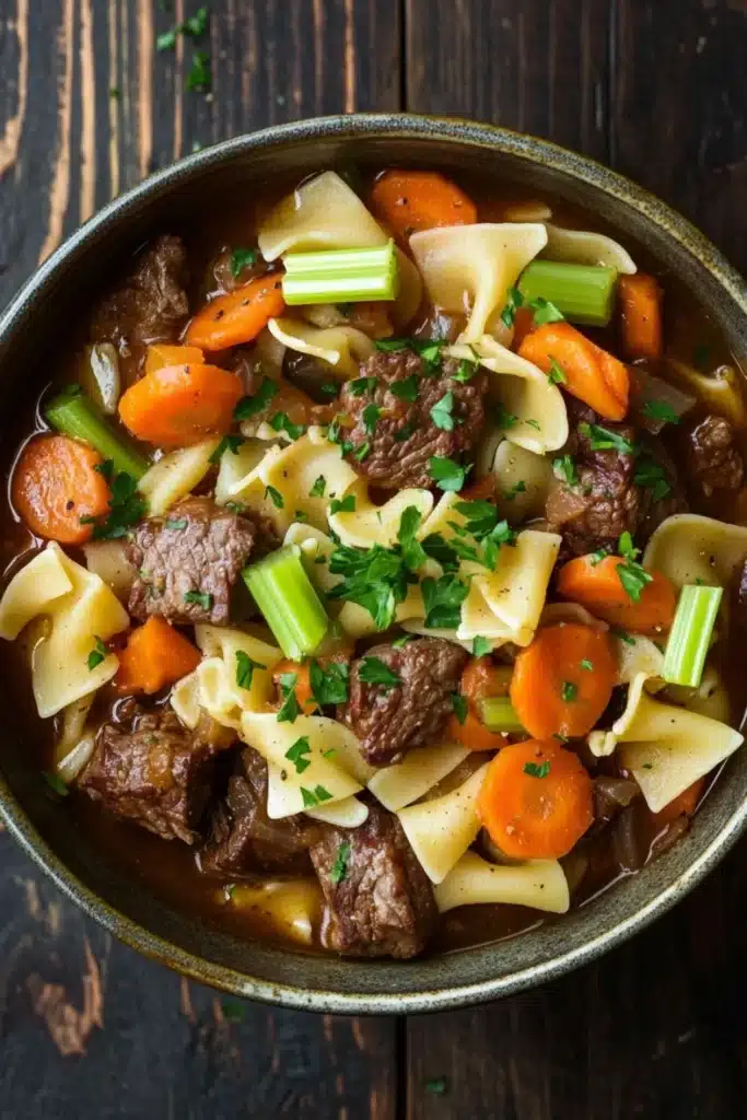Slow cooker stew meat and noodles - the image is a close-up of a bowl of beef noodle soup. the soup is made with chunks of beef, carrots, and celery, and is garnished with chopped parsley. the bowl is made of ceramic and is sitting on a dark wooden table. the broth is a rich brown color and appears to be thick and creamy. the noodles are long and thin, and the carrots are bright orange. the celery is a bright green color and is scattered throughout the soup, adding a pop of color to the dish.
