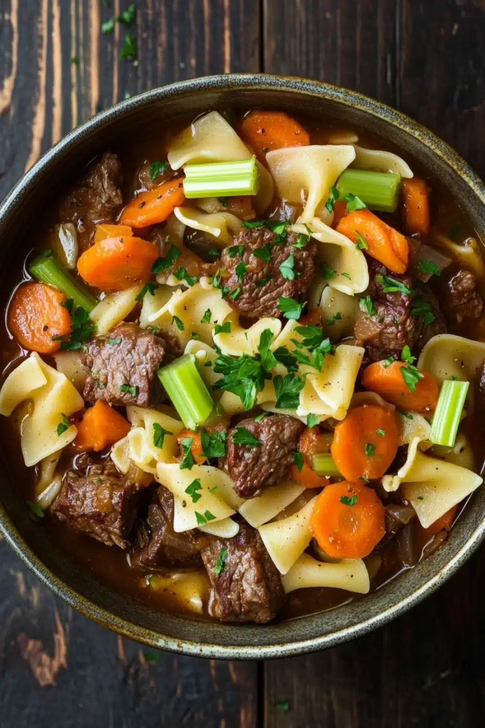 Slow cooker stew meat and noodles - the image is a close-up of a bowl of beef noodle soup. the soup is made with chunks of beef, carrots, and celery, and is garnished with chopped parsley. the bowl is made of ceramic and is sitting on a dark wooden table. the broth is a rich brown color and appears to be thick and creamy. the noodles are long and thin, and the carrots are bright orange. the celery is a bright green color and is scattered throughout the soup, adding a pop of color to the dish.