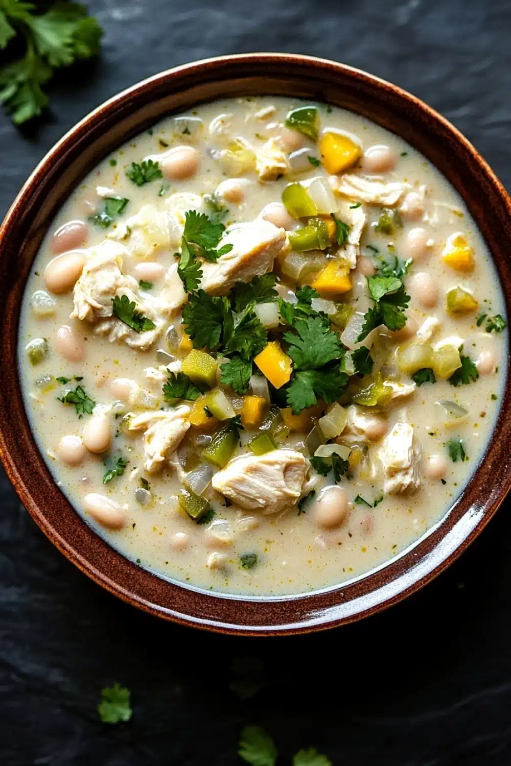 Slow cooker white chicken chili heavy cream - the image is a close-up of a bowl of soup. the soup appears to be a creamy white color with chunks of chicken and vegetables mixed in. the bowl is made of ceramic and has a brown rim. there are a few sprigs of fresh cilantro scattered around the bowl. the background is a dark grey countertop.