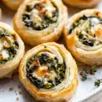 Spinach artichoke dip pinwheels - the image shows a plate of freshly baked spinach and feta puff pastry rolls. the rolls are golden brown and have a flaky texture. they are arranged in a circular pattern on a white plate with a sprig of thyme on the side. the spinach is a vibrant green color and appears to be cooked until soft and fluffy. the cheese is melted and bubbly on top, and there are small pieces of bacon scattered throughout the rolls.