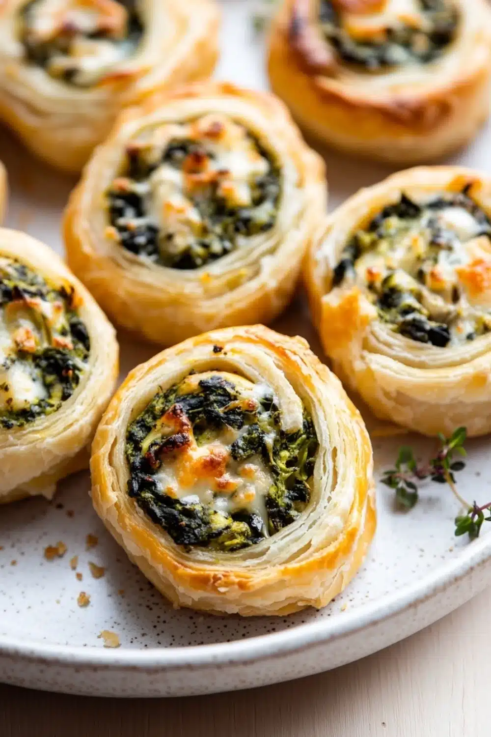 Spinach artichoke dip pinwheels - the image shows a plate of freshly baked spinach and feta puff pastry rolls. the rolls are golden brown and have a flaky texture. they are arranged in a circular pattern on a white plate with a sprig of thyme on the side. the spinach is a vibrant green color and appears to be cooked until soft and fluffy. the cheese is melted and bubbly on top, and there are small pieces of bacon scattered throughout the rolls.