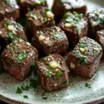 Steak bites with garlic butter stove top - the image shows a plate of square-shaped pieces of meat on a dark grey plate. the meat appears to be cooked medium-rare, with a dark brown color and a shiny texture. it is garnished with chopped herbs and chopped nuts, giving it a fresh and appetizing appearance. the plate is sitting on a wooden table, and the background is blurred, making the meat the focal point of the image.