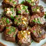 Steak bites with garlic butter stove top - the image shows a plate of cooked steak cubes. the steak cubes are arranged in a circular pattern on a light brown plate with a speckled pattern. they are covered in a dark brown sauce and garnished with chopped parsley and chopped nuts. the sauce appears to be drizzled over the top of the steak, giving it a glossy sheen. the plate is sitting on a white marble countertop.