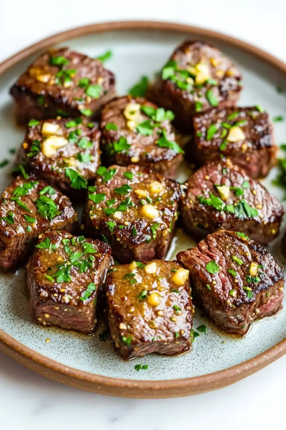 Steak bites with garlic butter stove top - the image shows a plate of cooked steak cubes. the steak cubes are arranged in a circular pattern on a light brown plate with a speckled pattern. they are covered in a dark brown sauce and garnished with chopped parsley and chopped nuts. the sauce appears to be drizzled over the top of the steak, giving it a glossy sheen. the plate is sitting on a white marble countertop.