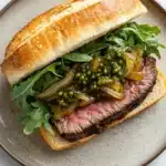 Steak sandwich with peppercorn sauce - the image shows a sandwich on a round plate with a white marble countertop. the sandwich is made with a toasted baguette and is filled with a generous portion of sliced steak, sliced onions, and a bed of green arugula. the steak appears to be cooked medium-rare and has a pinkish-brown crust. the onions are caramelized and appear to be seasoned with herbs and spices. the lettuce is a vibrant green color and is scattered throughout the sandwich. the plate is white with a speckled pattern.