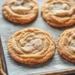 Sugar cookie chewy - the image shows a baking tray lined with freshly baked cookies. the cookies are round and golden brown in color, with a swirl pattern on top. they are arranged in a single layer on the tray, with some overlapping each other. the tray is placed on a wooden table, and the cookies appear to be freshly baked. the background is blurred, but it appears to be a kitchen countertop.