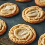 Sugar cookie chewy - the image shows a baking tray with freshly baked cookies on it. the cookies are round and golden brown in color, with a swirl pattern on top. they are arranged in a single layer on the tray, with some overlapping each other. the tray is lined with parchment paper, and there is a metal spatula resting on the side. the background is dark, making the cookies stand out.