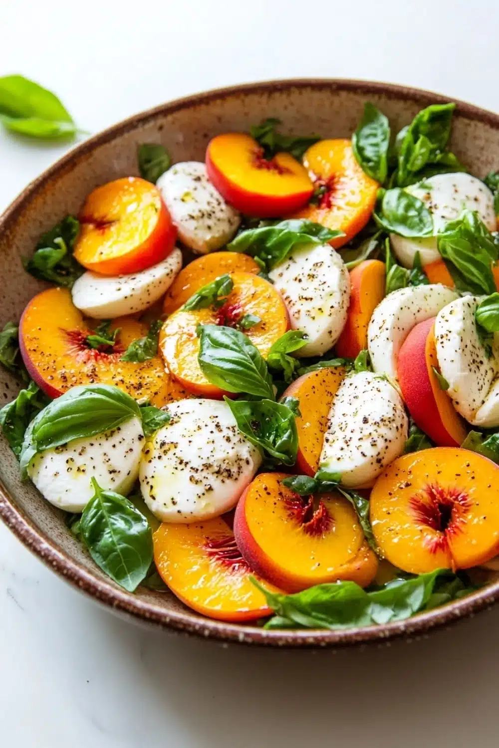 Summer peach caprese salad - the image is a close-up of a salad in a bowl. the salad is made up of sliced peaches, mozzarella cheese, and fresh basil leaves. the peaches are arranged in a circular pattern, with some overlapping each other. the cheese is white and appears to be soft and fluffy. the basil leaves are a vibrant green color and are scattered throughout the salad. the bowl is a light brown color and is sitting on a white marble countertop. there are a few sprigs of fresh basil scattered around the bowl, adding a pop of color to the dish.
