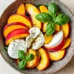 Summer peach caprese salad - the image is a close-up of a bowl of salad. the bowl is made of a light-colored ceramic material and is placed on a beige-colored tablecloth. the salad is made up of sliced peaches, sliced oranges, and basil leaves. on top of the peaches and oranges, there is a dollop of mozzarella cheese. the peaches are arranged in a circular pattern, with some overlapping each other. the basil leaves are scattered throughout the salad, adding a pop of green color to the dish. the overall presentation of the salad is fresh and appetizing.