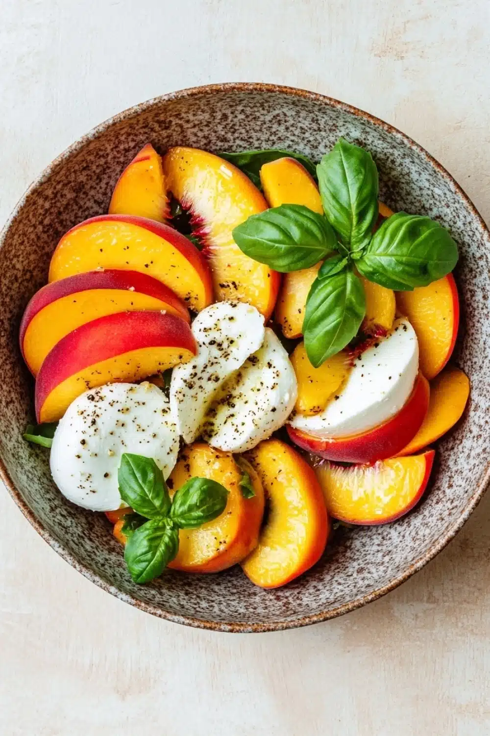 Summer peach caprese salad - the image is a close-up of a bowl of salad. the bowl is made of a light-colored ceramic material and is placed on a beige-colored tablecloth. the salad is made up of sliced peaches, sliced oranges, and basil leaves. on top of the peaches and oranges, there is a dollop of mozzarella cheese. the peaches are arranged in a circular pattern, with some overlapping each other. the basil leaves are scattered throughout the salad, adding a pop of green color to the dish. the overall presentation of the salad is fresh and appetizing.