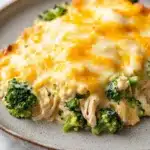 Trish yearwood chicken broccoli casserole - the image shows a plate of food with a casserole dish on it. the dish appears to be made with chicken and broccoli, and it is covered in melted cheese. the cheese is a bright yellow color and is spread evenly over the top of the dish. the broccoli is a vibrant green color and looks fresh and healthy. the plate is a light brown color with a speckled pattern around the edges. the background is blurred, but it seems to be a kitchen countertop.