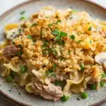 Tuna casserole no cheese - the image is a close-up of a plate of pasta dish. the dish appears to be a type of fettuccine alfredo, with a creamy white sauce and chunks of meat and vegetables mixed in. the pasta is cooked al dente and is topped with a crumbled breadcrumb topping. there are also small pieces of sliced mushrooms scattered throughout the dish, adding a pop of green color. the plate is sitting on a white marble countertop, and there are a few sprigs of parsley scattered around the plate.