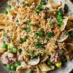 Tuna casserole no cheese - the image is a close-up of a bowl of pasta dish. the dish appears to be a type of fettuccine alfredo pasta, with chunks of meat and vegetables mixed in. the pasta is cooked al dente and is topped with a crumbled breadcrumb topping. the vegetables include peas, mushrooms, and corn kernels. the bowl is sitting on a wooden table with a green napkin and a sprig of parsley scattered around it. the overall color scheme of the dish is warm and inviting.
