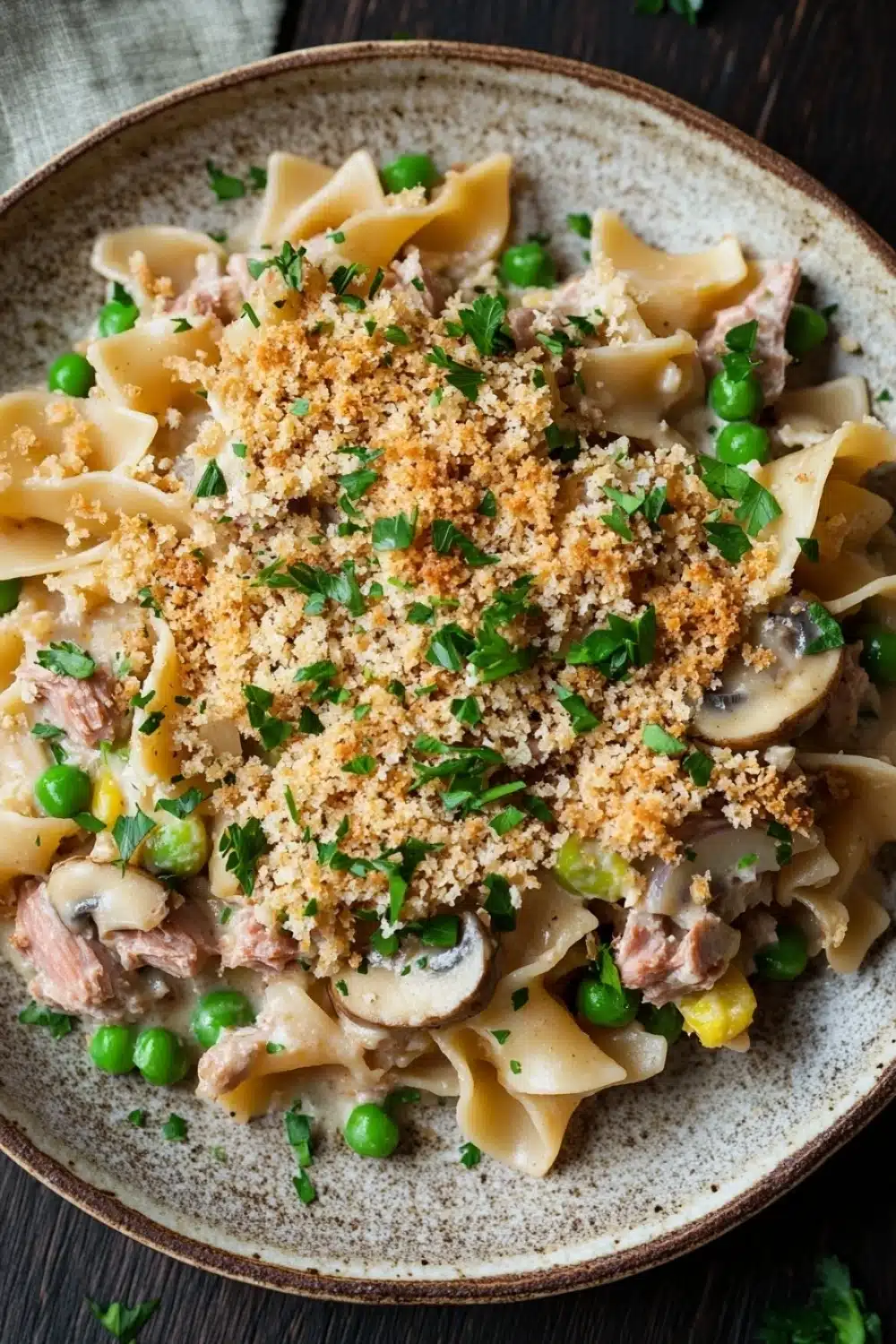 Tuna casserole no cheese - the image is a close-up of a bowl of pasta dish. the dish appears to be a type of fettuccine alfredo pasta, with chunks of meat and vegetables mixed in. the pasta is cooked al dente and is topped with a crumbled breadcrumb topping. the vegetables include peas, mushrooms, and corn kernels. the bowl is sitting on a wooden table with a green napkin and a sprig of parsley scattered around it. the overall color scheme of the dish is warm and inviting.