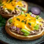 Tuna melt bagel - the image shows two sandwiches on a blue plate. the sandwiches are made with a toasted bun and are topped with a generous amount of melted cheese. the cheese is a bright yellow color and is drizzled over the top of the sandwiches. there are also chopped green onions and red onions scattered on top. the plate is garnished with a sprig of parsley. the background is dark and out of focus, making the sandwiches stand out.