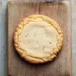 Vanilla bean shortbread cookie - the image shows a round cookie on a wooden cutting board. the cookie appears to be freshly baked and has a golden brown color. it has a crumbly texture and is covered in small black speckles. the cutting board is square in shape and is placed on a weathered wooden surface. the background is blurred, making the cookie the focal point of the image.
