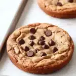 Vegan peanut butter cookie cake - the image shows two freshly baked chocolate chip cookies on a baking tray. the cookies are round and golden brown in color, with chocolate chips scattered throughout. they are placed on a white marble countertop, and there is a pair of chopsticks resting on the tray next to them. the background is blurred, but it appears to be a kitchen countertop.
