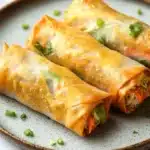 Vegetarian spring rolls air fryer - the image shows three spring rolls on a gray plate. the spring rolls are golden brown and appear to be freshly cooked. they are filled with a mixture of vegetables, including carrots, celery, and onions, and are garnished with chopped parsley. the plate is sitting on a white marble countertop.