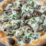White sauce truffle pizza - the image is of a freshly baked pizza on a wooden cutting board. the pizza has a golden brown crust and is topped with melted white cheese and sliced mushrooms. the mushrooms are arranged in a circular pattern on top of the cheese, with some overlapping each other. there are also some chopped herbs scattered around the pizza, adding a pop of green color to the dish. the background is a dark grey countertop, and there is a small sprig of parsley on the side.