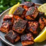 Air fryer salmon bites blackened - the image shows a plate of grilled salmon on a dark grey plate. the salmon is cooked to a golden brown color and has a crispy texture. it is garnished with two lemon wedges on either side of the plate, adding a pop of color to the dish. in the background, there is a bowl of fresh parsley and a sprig of parsley. the plate is sitting on a black countertop.
