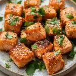 Air fryer salmon bites honey siracha - the image shows a plate of cooked salmon cubes. the salmon cubes are golden brown and have sesame seeds sprinkled on top. they are arranged in a circular pattern on the plate, with some overlapping each other. the plate is white with a light blue rim and is garnished with fresh cilantro leaves. the background is blurred, but it appears to be a wooden table.