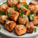 Air fryer salmon bites honey siracha - the image shows a plate of grilled salmon with sesame seeds on top. the salmon is golden brown and appears to be seasoned with herbs and spices. the plate is white with a speckled pattern and is garnished with fresh cilantro leaves. the background is a white marble countertop.