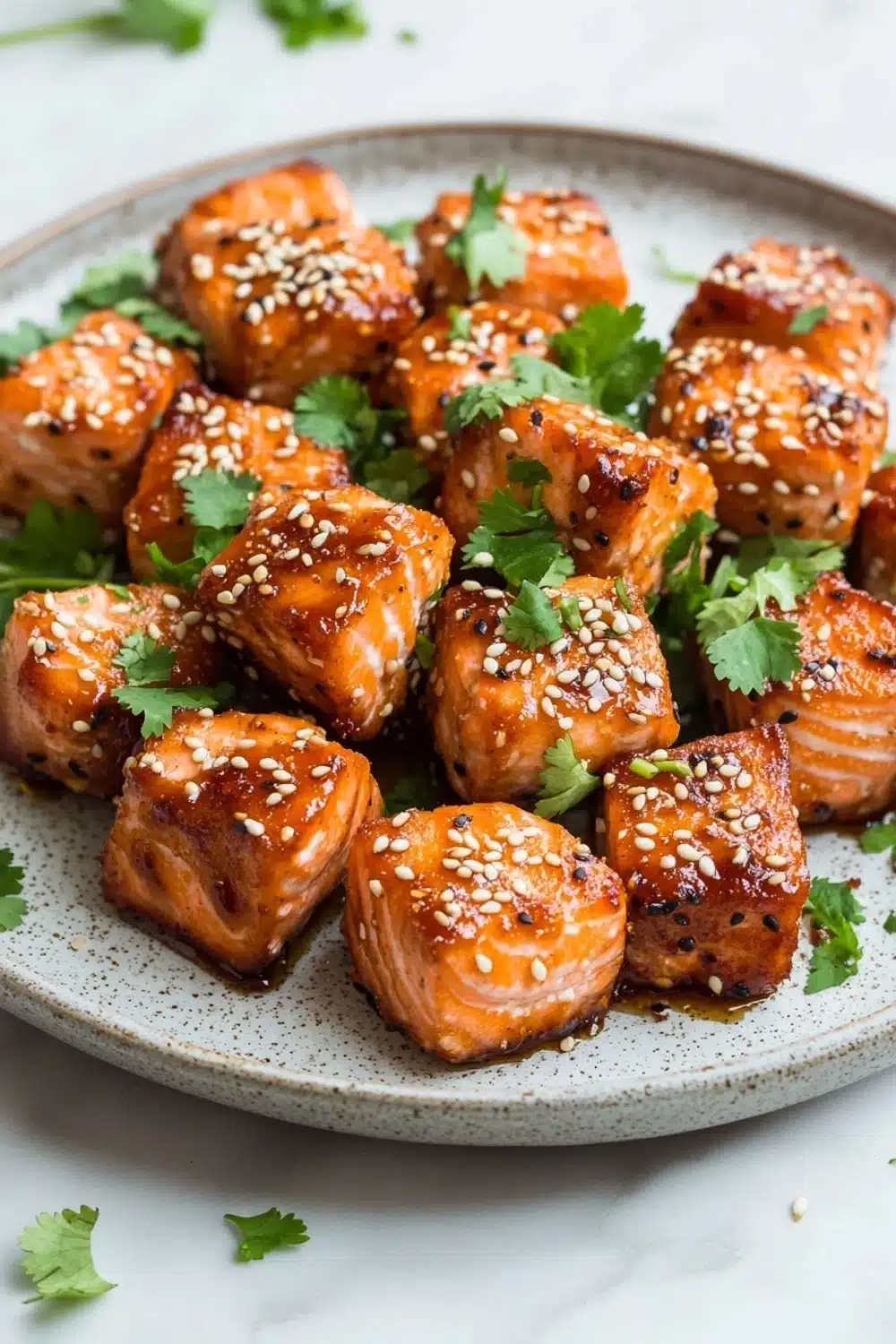 Air fryer salmon bites honey siracha - the image shows a plate of grilled salmon with sesame seeds on top. the salmon is golden brown and appears to be seasoned with herbs and spices. the plate is white with a speckled pattern and is garnished with fresh cilantro leaves. the background is a white marble countertop.