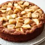 Apple cake no flour - the image is of a freshly baked apple tart on a white plate. the tart has a golden brown crust and is topped with sliced apples arranged in a circular pattern. the apples are cut into small cubes and are evenly spaced along the top of the tart. the plate has a speckled pattern around the edges. the background is blurred, but it appears to be a kitchen countertop.