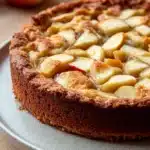 Apple cake with apple pie filling - the image is of a freshly baked apple tart on a white plate. the tart has a golden brown crust on top and is filled with sliced apples arranged in a criss-cross pattern. the apples are cut into small cubes and appear to be freshly baked. in the background, there are two whole apples and a few more apples on a wooden table. the overall color scheme of the image is warm and inviting.