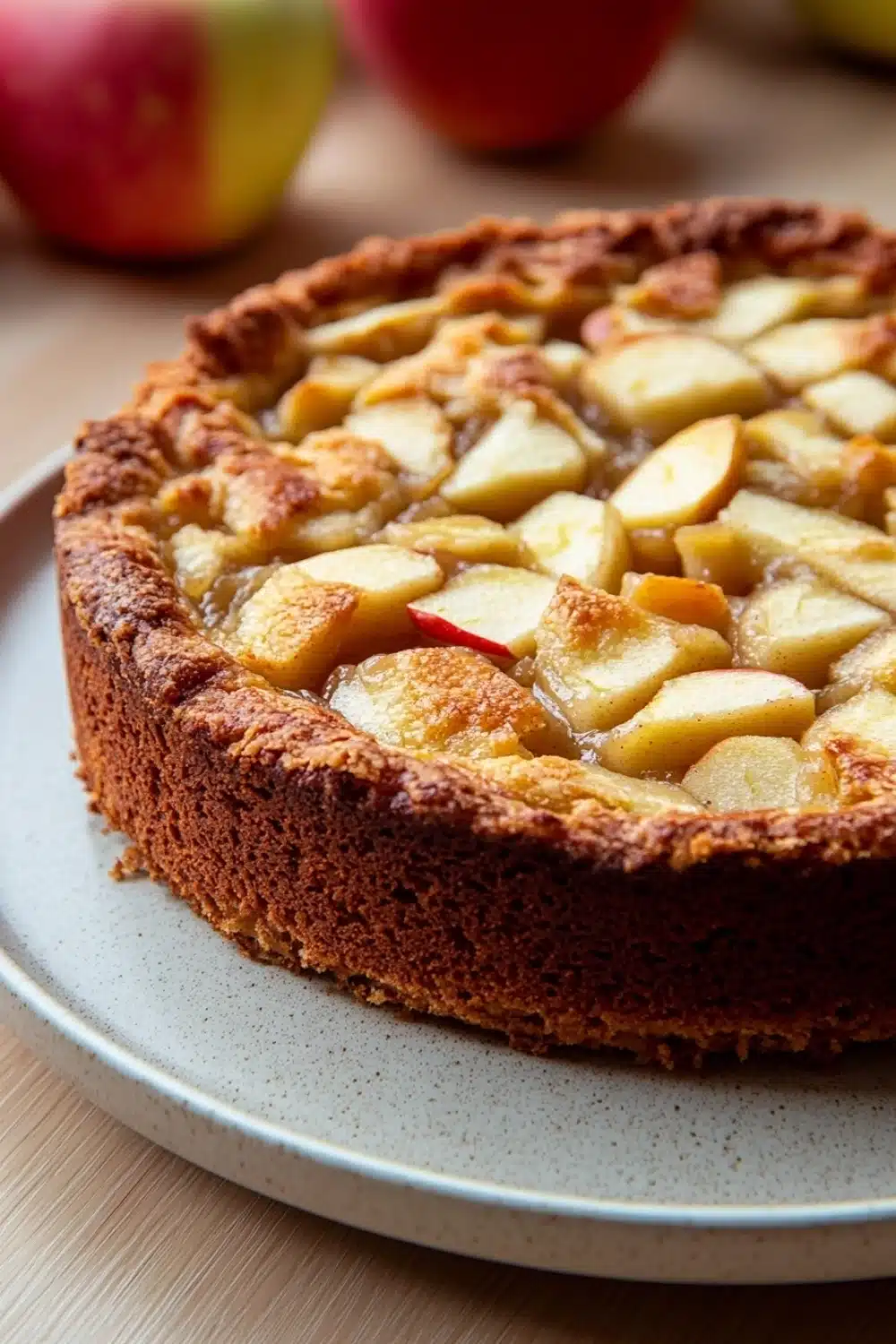 Apple cake with apple pie filling - the image is of a freshly baked apple tart on a white plate. the tart has a golden brown crust on top and is filled with sliced apples arranged in a criss-cross pattern. the apples are cut into small cubes and appear to be freshly baked. in the background, there are two whole apples and a few more apples on a wooden table. the overall color scheme of the image is warm and inviting.