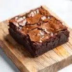 Banana bread brownies - the image shows a rectangular piece of chocolate cake on a wooden cutting board. the cake appears to be freshly baked and has a rich, dark brown color. the top of the cake is covered in a layer of chocolate frosting, which is drizzled with a light brown glaze. the cutting board is placed on a white marble countertop. the background is blurred, so the focus is on the cake.