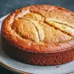 Banana cake in air fryer - the image is a close-up of a round cake on a white plate. the cake appears to be freshly baked and has a golden brown crust on top. it has a criss-cross pattern on the sides, with the top half of the cake being slightly darker than the bottom half. the plate is sitting on a dark grey countertop. the background is blurred, making the cake the focal point of the image.