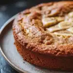 Banana cake in air fryer - the image is a close-up of a round cake on a silver plate. the cake appears to be freshly baked and has a golden brown crust on top. on top of the cake, there are sliced bananas arranged in a circular pattern. the bananas are cut into thin slices and are scattered across the cake. the plate is sitting on a dark grey countertop. the background is blurred, making the cake the focal point of the image.