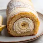 Banana cake roll - the image shows a slice of banana roll on a white plate. the roll is golden brown in color and has a crumbly texture. it is dusted with powdered sugar, giving it a shiny appearance. the center of the roll is filled with a creamy white filling, which appears to be a banana peel. on the left side of the plate, there is a whole banana, and on the right side, there are two whole bananas. the background is a plain white surface.