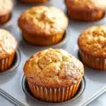 Banana muffin pictures - the image shows a muffin tin filled with freshly baked muffins. the muffins are golden brown in color and have a crumbly texture on top. they are arranged in a single layer in the tin, with some overlapping each other. the tin is placed on a white marble countertop. the background is blurred, but it appears to be a kitchen countertop with more muffins scattered around.