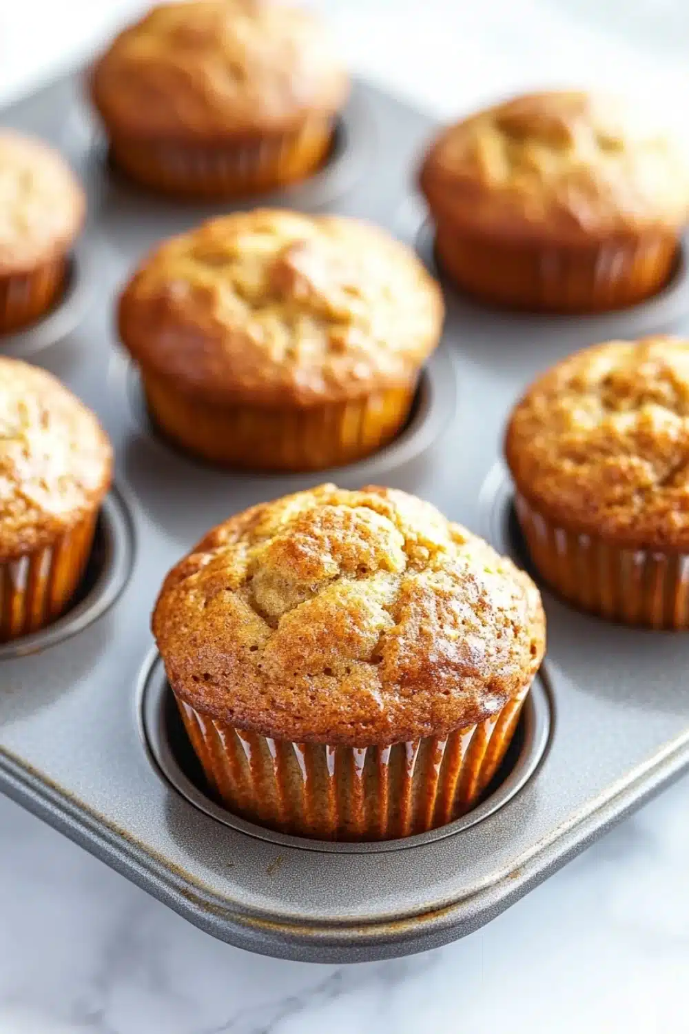 Banana muffin pictures - the image shows a muffin tin filled with freshly baked muffins. the muffins are golden brown in color and have a crumbly texture on top. they are arranged in a single layer in the tin, with some overlapping each other. the tin is placed on a white marble countertop. the background is blurred, but it appears to be a kitchen countertop with more muffins scattered around.