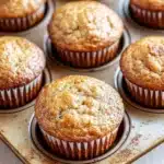 Banana muffin pictures - the image shows a muffin tin filled with freshly baked muffins. the muffins are golden brown in color and have a crumbly texture on top. they are arranged in a circular pattern in the tin, with some overlapping each other. the tin is sitting on a white wooden surface. the background is blurred, but it appears to be a kitchen countertop.