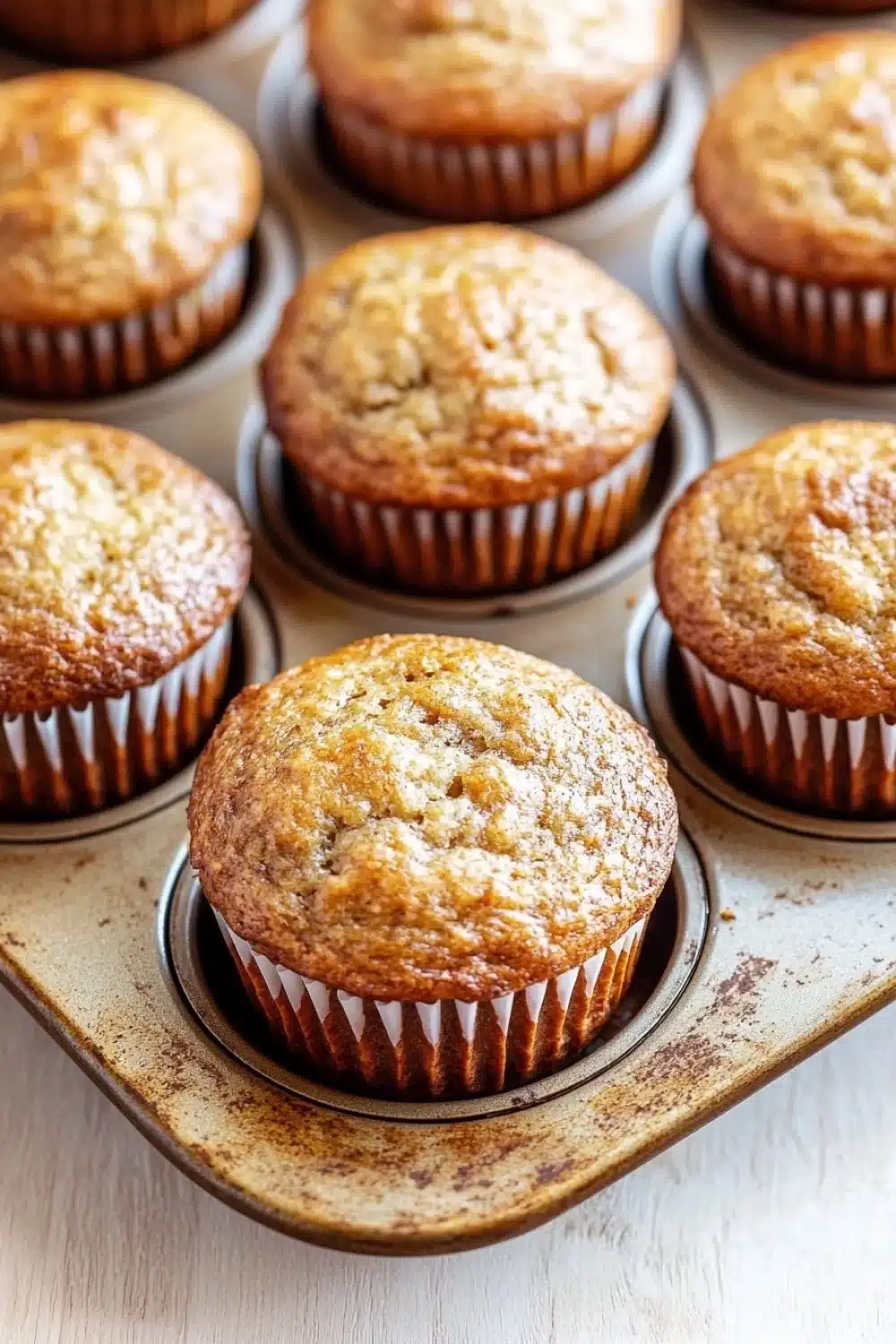 Banana muffin pictures - the image shows a muffin tin filled with freshly baked muffins. the muffins are golden brown in color and have a crumbly texture on top. they are arranged in a circular pattern in the tin, with some overlapping each other. the tin is sitting on a white wooden surface. the background is blurred, but it appears to be a kitchen countertop.