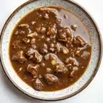 Beef chicken gravy - the image is a close-up of a bowl of beef stew. the bowl is white with a brown rim and is placed on a white marble countertop. the stew is a rich, dark brown color and appears to be thick and thick, with chunks of beef and vegetables visible in the broth. the beef is cooked until tender and tender, and there are small pieces of onions and celery scattered throughout the dish. the dish looks appetizing and ready to eat.
