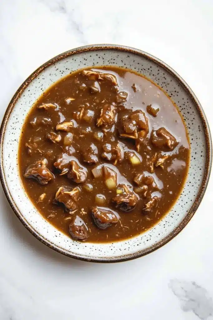 Beef chicken gravy - the image is a close-up of a bowl of beef stew. the bowl is white with a brown rim and is placed on a white marble countertop. the stew is a rich, dark brown color and appears to be thick and thick, with chunks of beef and vegetables visible in the broth. the beef is cooked until tender and tender, and there are small pieces of onions and celery scattered throughout the dish. the dish looks appetizing and ready to eat.