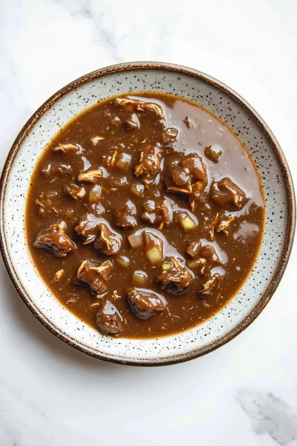 Beef chicken gravy - the image is a close-up of a bowl of beef stew. the bowl is white with a brown rim and is placed on a white marble countertop. the stew is a rich, dark brown color and appears to be thick and thick, with chunks of beef and vegetables visible in the broth. the beef is cooked until tender and tender, and there are small pieces of onions and celery scattered throughout the dish. the dish looks appetizing and ready to eat.
