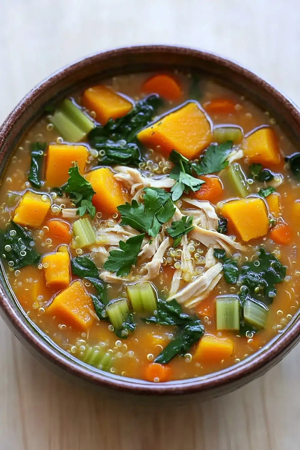 Butternut squash chicken quinoa soup - the image is a close-up of a bowl of soup. the soup appears to be a creamy orange color with chunks of orange and green vegetables mixed in. there are also chunks of white chicken and green onions scattered throughout the soup. on top of the soup, there is a sprig of fresh cilantro. the bowl is made of ceramic and is sitting on a wooden table. the background is blurred, making the soup the focal point of the image.