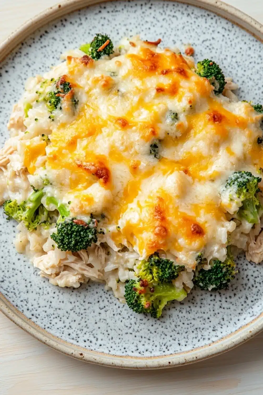 Cheesy chicken rice broccoli casserole - the image is a close-up of a plate of food. the plate is round and has a blue and white speckled pattern. on top of the plate, there is a layer of creamy white rice with chunks of chicken and broccoli scattered throughout. the chicken appears to be cooked and has melted cheese on top. the broccoli is bright green and looks fresh and vibrant. the dish looks appetizing and ready to eat.