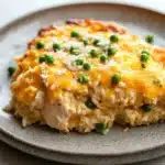 Chicken rice casserole crockpot - the image shows a plate of food on a white tablecloth. the plate is round and has a light brown color with a speckled pattern. on top of the plate, there is a serving of risotto with a layer of melted cheese on top. the cheese is a bright yellow color and appears to be melted and bubbly. there are small pieces of green peas scattered on top, adding a pop of color to the dish. the risotto is a creamy white color and looks fluffy and fluffy.