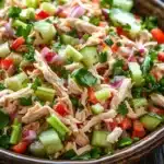 Chicken salad no mayo greek yogurt - the image is a close-up of a large bowl filled with a colorful salad. the salad appears to be made up of shredded chicken, sliced cucumbers, red onions, and green onions. the chicken is cut into small pieces and is mixed with a variety of vegetables, including red bell peppers, green celery, and red onions. there are also some cilantro leaves scattered throughout the salad, adding a pop of green color to the dish. the bowl is sitting on a dark wooden table with a gray cloth napkin on the side. the background is blurred, but it seems to be a kitchen countertop.