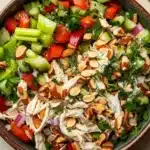 Chicken salad no mayo greek yogurt - the image is of a large bowl filled with a colorful salad. the salad is made up of various ingredients such as sliced cucumbers, red bell peppers, sliced onions, sliced almonds, and dill. the ingredients are mixed together in a creamy dressing, which appears to be chicken or chicken salad. there are also some fresh herbs and spices scattered throughout the salad, adding a pop of green color to the dish. the bowl is sitting on a wooden table, and there is a sprig of parsley on the side.